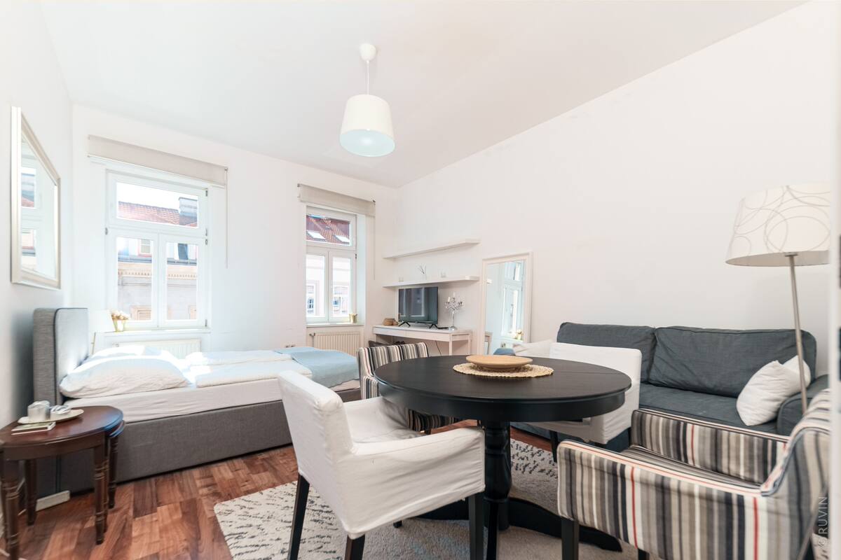 A modern, well-lit room with a bed, a sofa, a dining table, and a kitchen area.