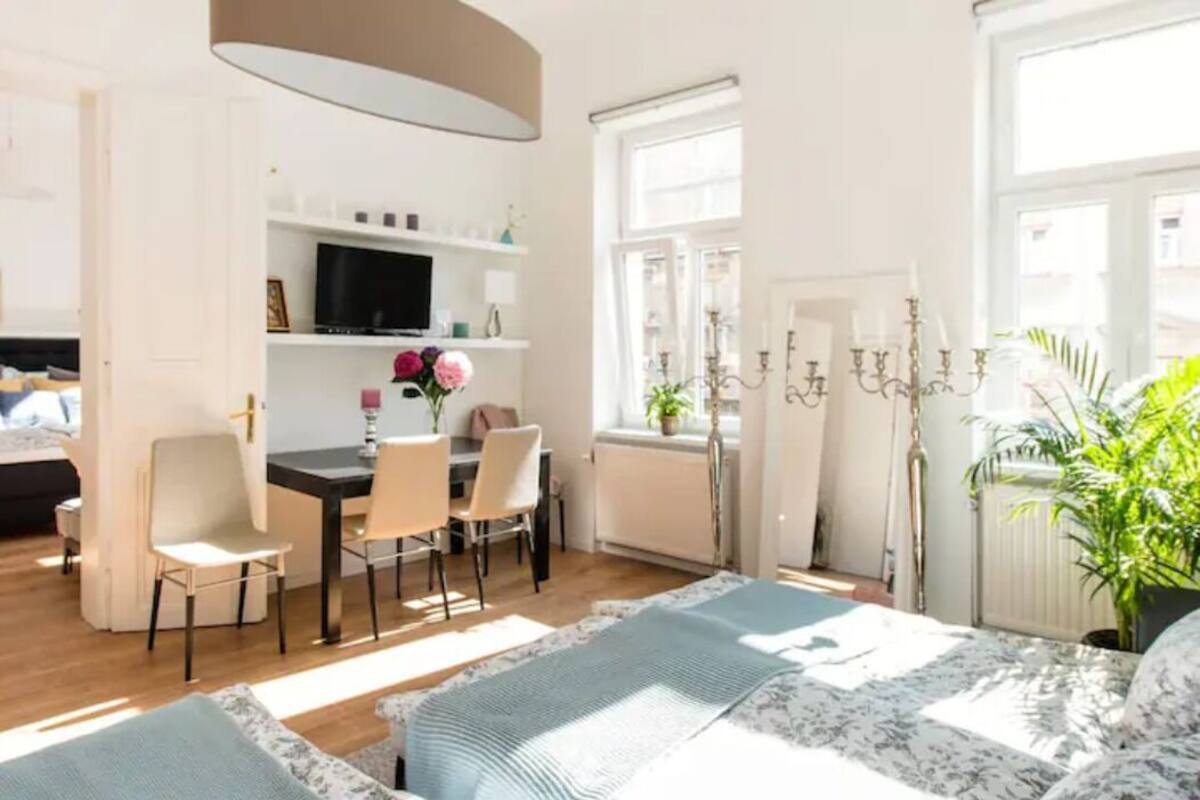 A bright, well-lit room with a bed, a desk, and chairs. There is a TV on the desk, a window with curtains, and a radiator.