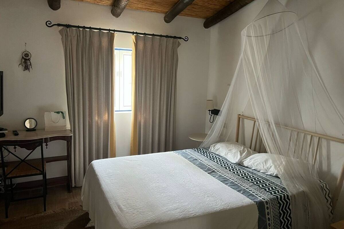 A bedroom with a bed, a desk, a chair, a bedside table, and a window with curtains.