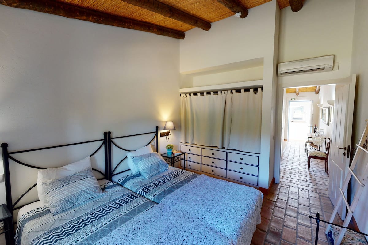 A bedroom with a bed, bedside tables, a lamp, a mirror, and an air conditioning unit.
