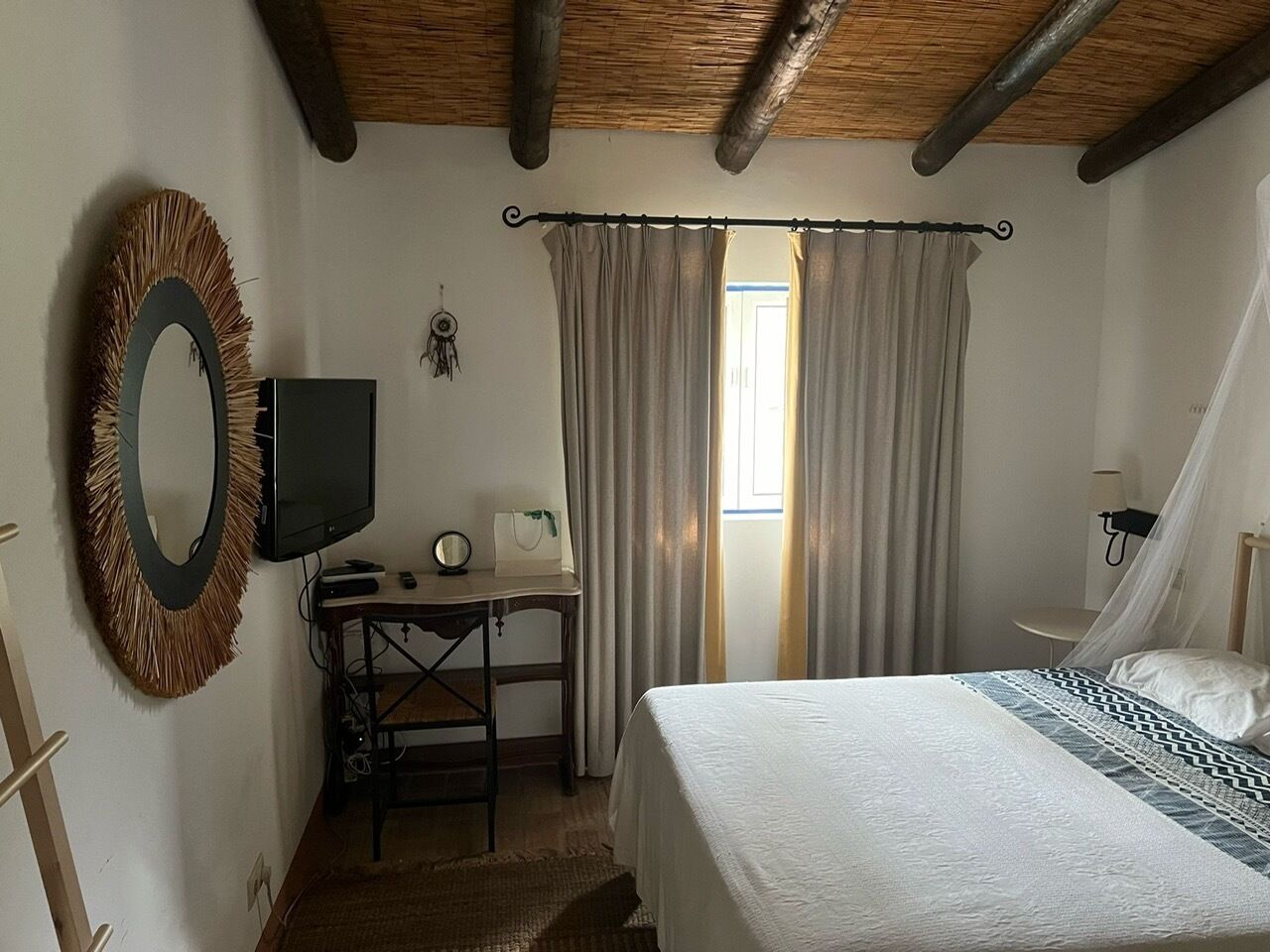 Ein Schlafzimmer mit einem Bett, einem Schreibtisch mit Fernseher, einem Spiegel und Vorhängen.