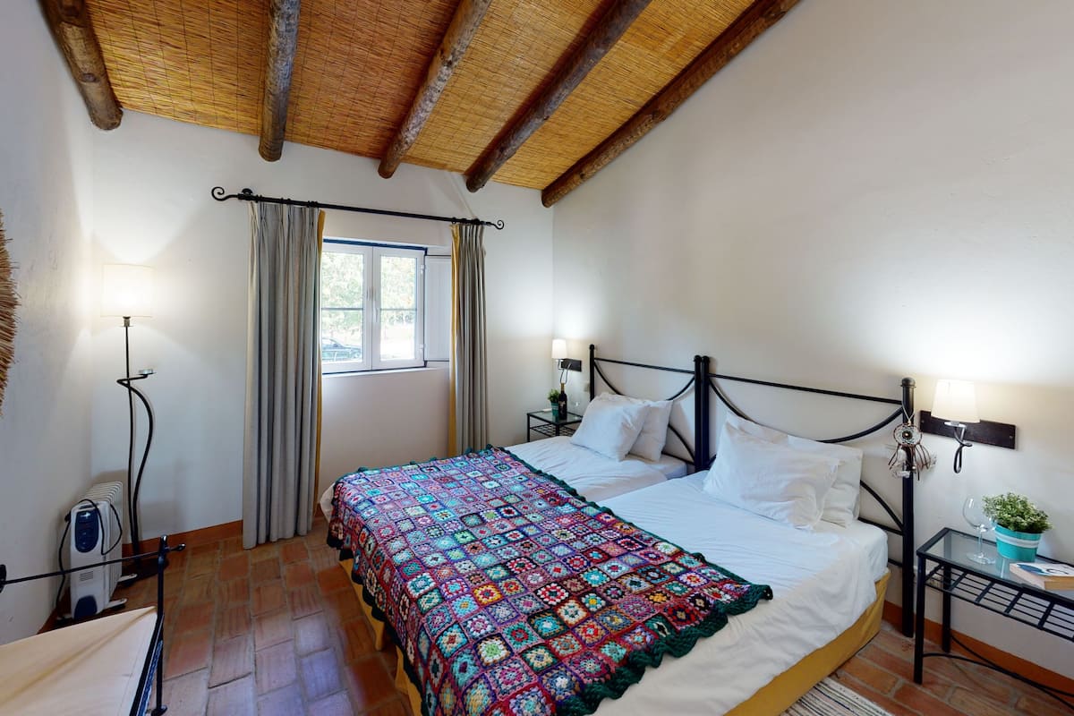 A bedroom with a large bed, a bedside table with a lamp, a window with curtains, a mirror, and a ceiling with exposed beams.