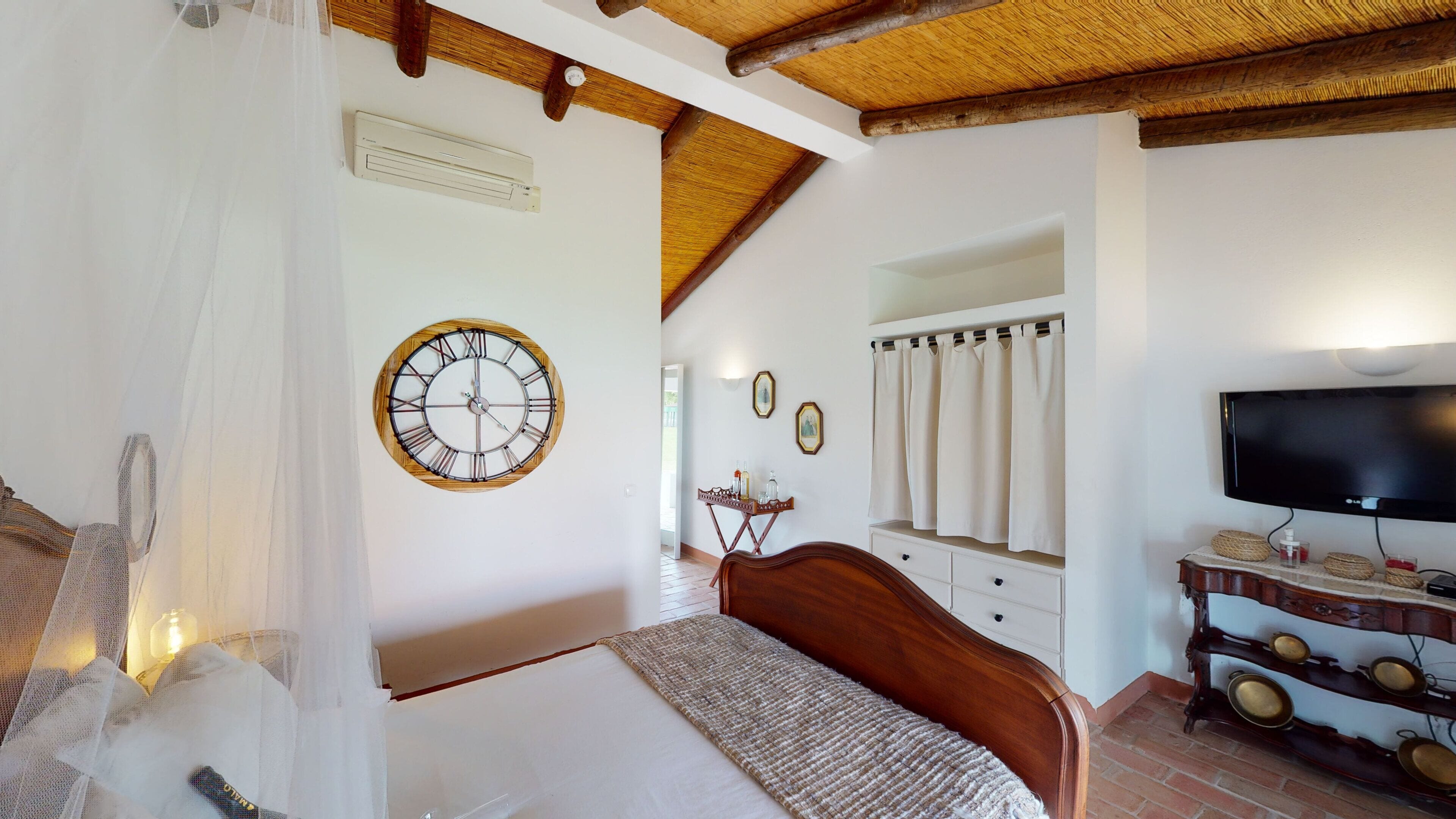 Un dormitorio con cama, techo de madera, televisor montado en la pared, mesita de noche y espejo.