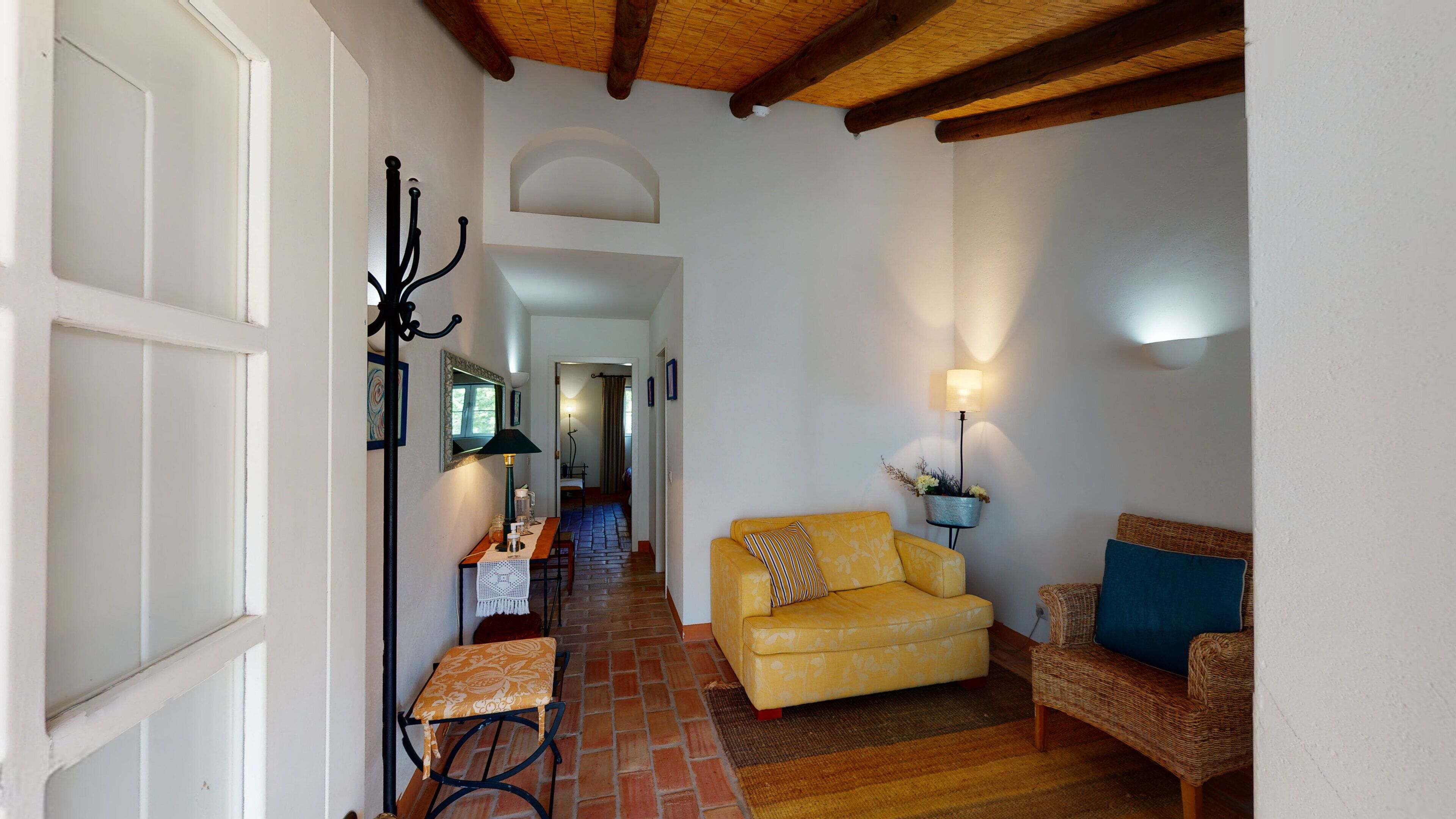 Un couloir avec un canapé jaune, un fauteuil en osier et une table avec une lampe.