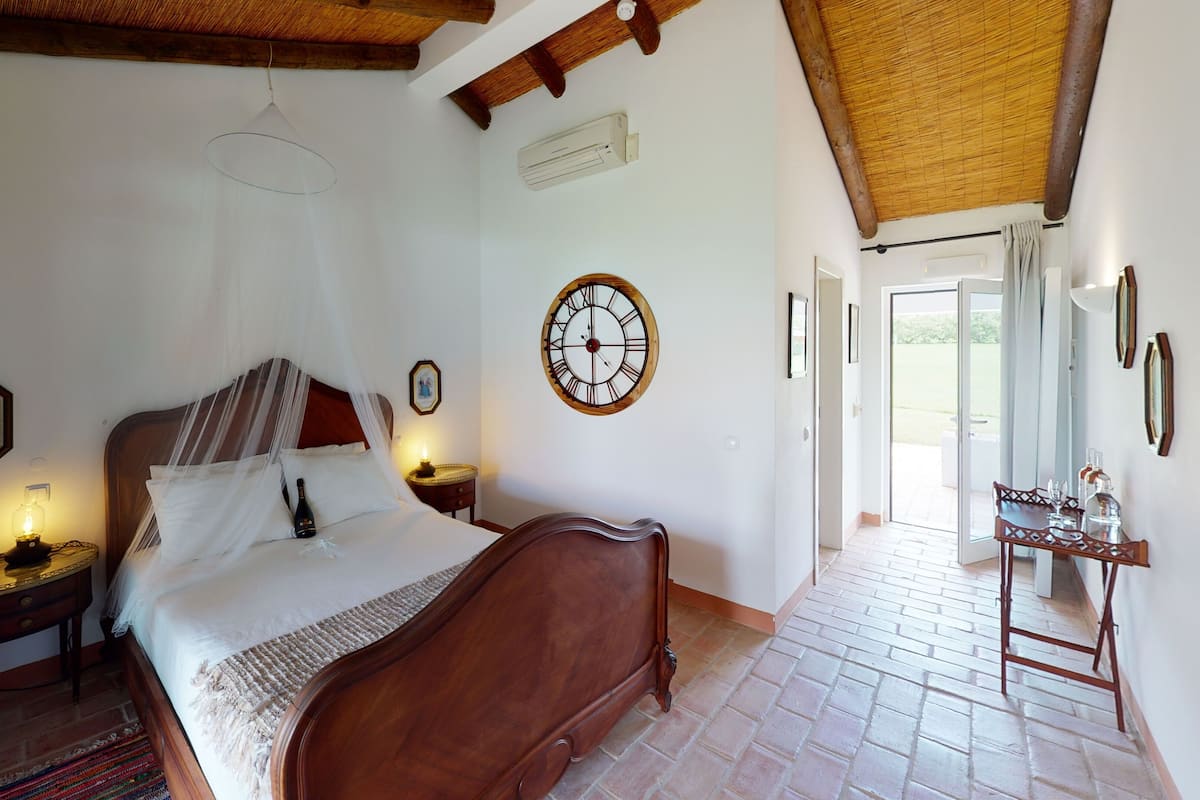 A bedroom with a wooden bed, a bedside table with a lamp, a wall clock, and a window leading to a balcony.