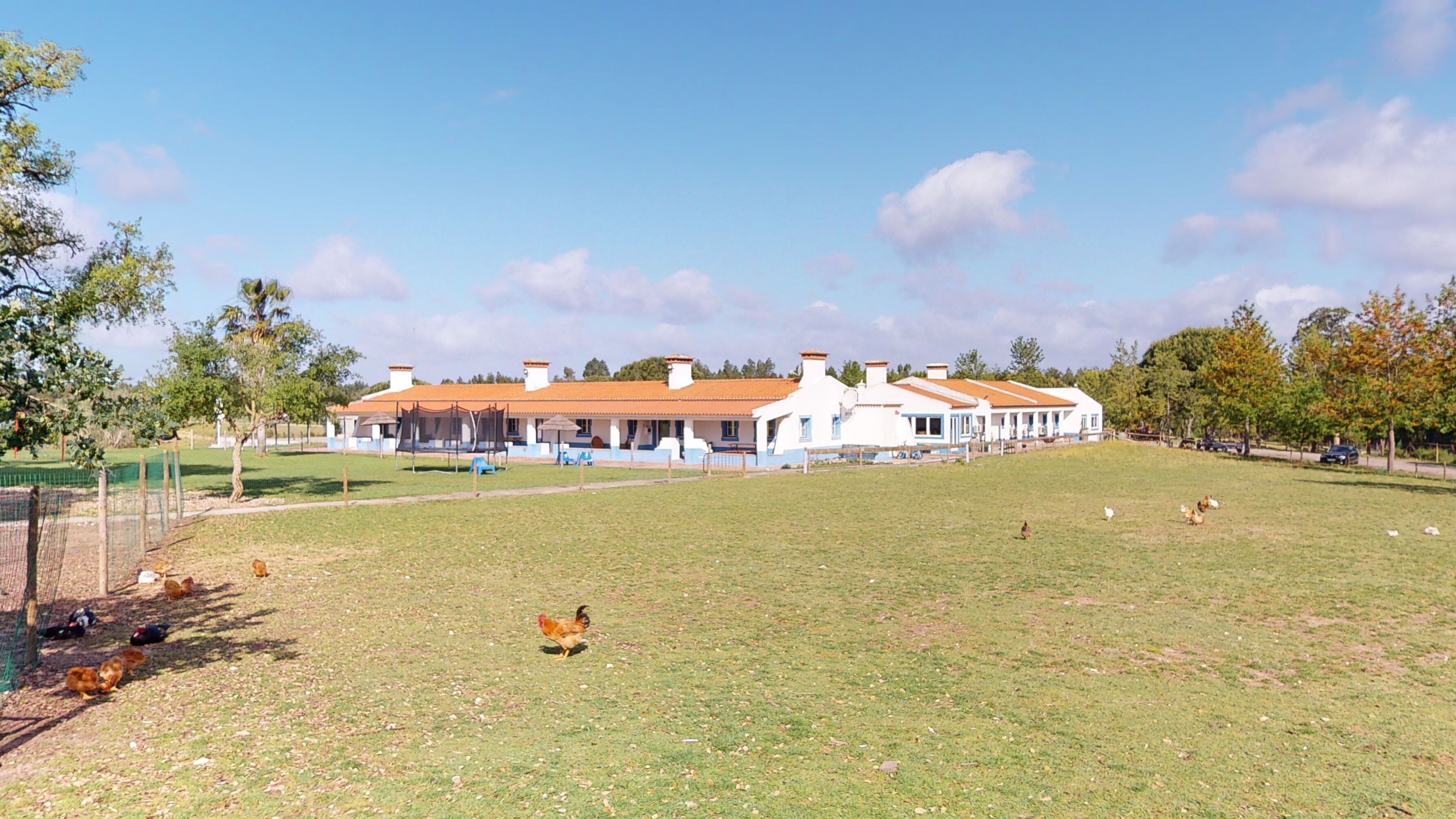 A large, single-story building with a red-tiled roof and white walls, surrounded by a grassy field.