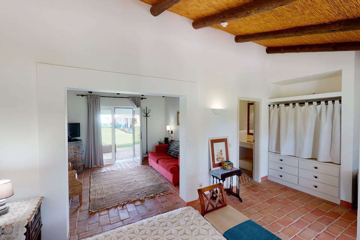 A spacious room with a red sofa, a small table with a lamp, a white wardrobe, and a large window with curtains.
