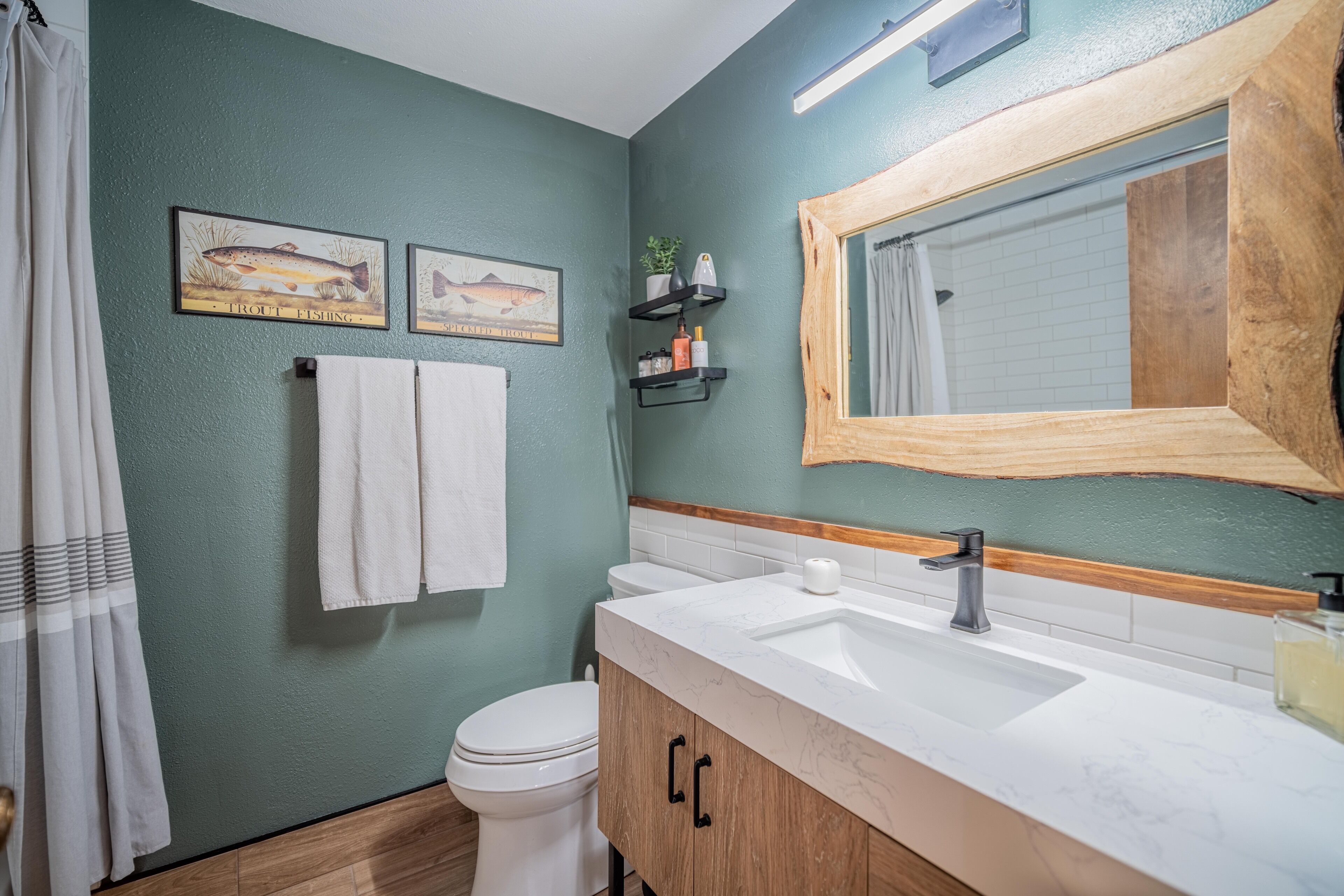 Une salle de bain avec un mur vert, des accessoires blancs, un meuble en bois et deux tableaux représentant des poissons.