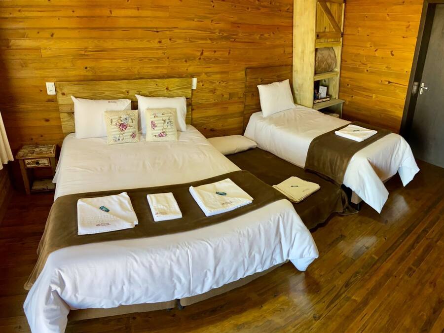 Una habitación tipo cabaña de madera con dos camas, cada una con sábanas blancas y almohadones florales. Tiene pisos de madera y cabecera de madera.