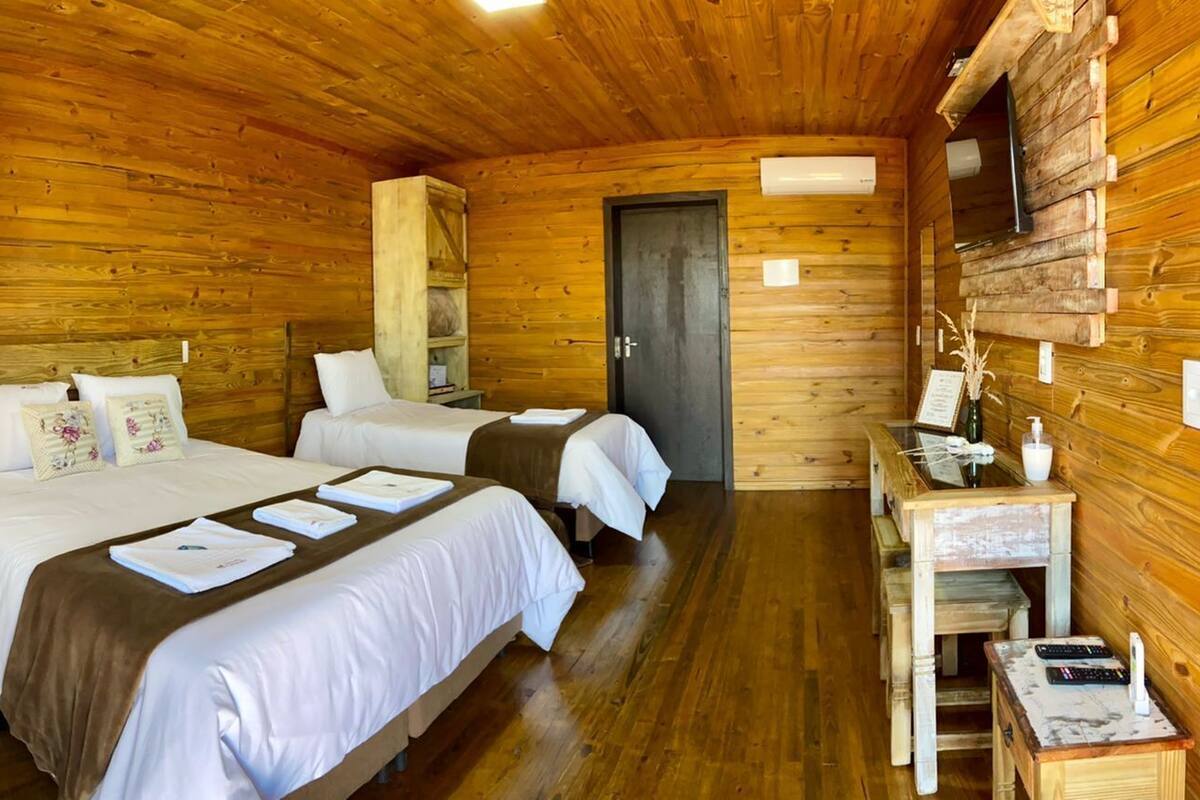 Quarto de cabana de madeira com duas camas, uma mesinha e uma prateleira.