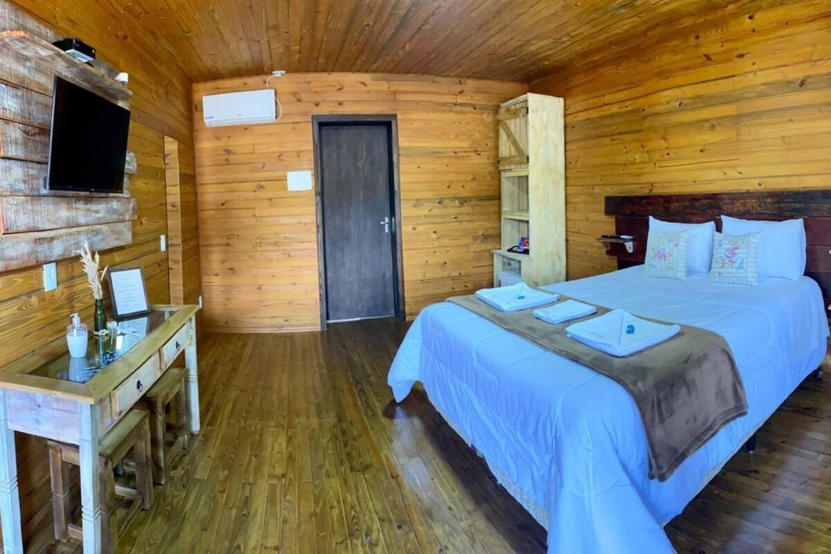Quarto de cabana de madeira com uma cama, uma TV fixada na parede, uma mesa pequena e uma porta.
