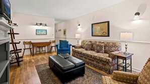 A living room with a sofa, ottoman, and chairs, a TV, and artwork on the walls. - GC106•walk downtown•onsite pool•private hot tub (Gatlinburg)