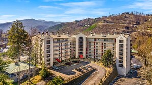 A multi-story hotel with a central courtyard, parking area, and a mountainous backdrop. - GC507•walk downtown •onsite heated pool (Gatlinburg)