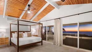 Ein Schlafzimmer mit Himmelbett, Deckenventilator und einer großen Schiebetür, die auf einen Balkon mit Sonnenuntergangsblick führt.