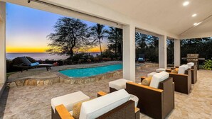 Ein überdachter Außenbereich mit Pool, Korbmöbeln und Blick auf den Sonnenuntergang über dem Meer.