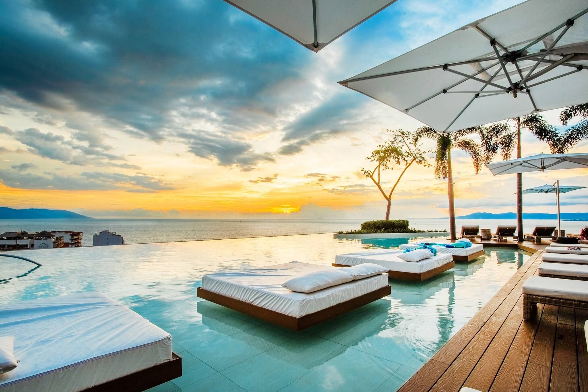 Une piscine à débordement avec des chaises longues et des parasols, offrant une vue imprenable sur le coucher de soleil sur l’océan.