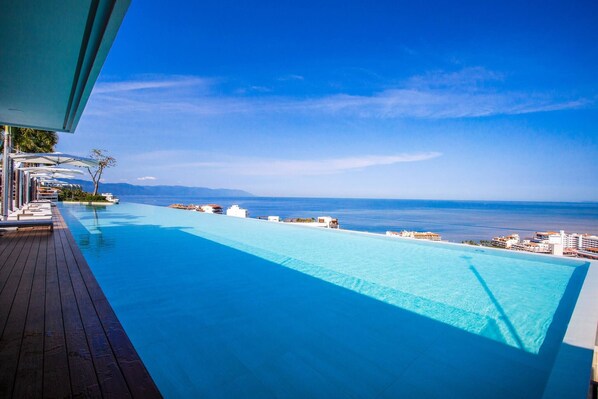 Ein langer, klarer, blauer Infinity-Pool mit einer Holzterrasse auf einer Seite und Blick auf den Ozean sowie entfernte Berge.