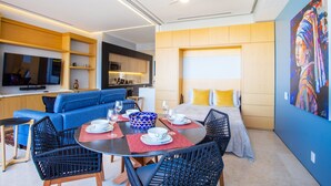 A modern hotel room with a dining area, a sofa, a bed, and a painting on the wall.
