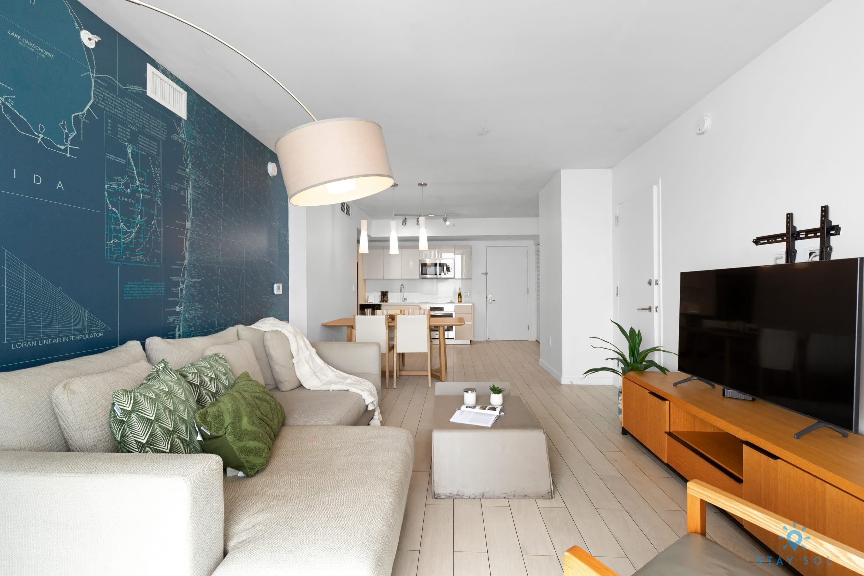 A modern living room with a sofa, ottoman, and a TV on a wooden stand. A kitchen is visible in the background.