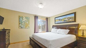 Une chambre à coucher comprenant un lit, une commode, une table de chevet, une lampe, un tableau et une fenêtre avec des rideaux.
