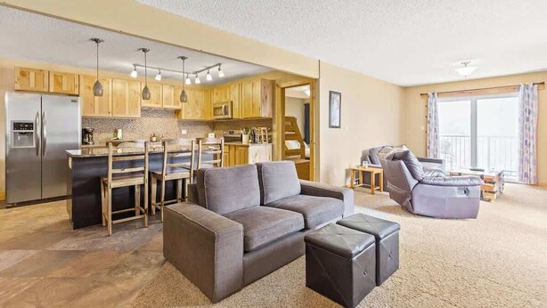 Un salon moderne comprenant un canapé, un pouf et un coin repas avec des meubles de rangement en bois et un réfrigérateur.