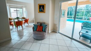 Ein großzügiger Wohnbereich mit Esstisch und Stühlen, eine gemütliche Sitzecke mit Kissen, einem Gemälde an der Wand und Blick auf einen Swimmingpool durch große Glasfenster.
