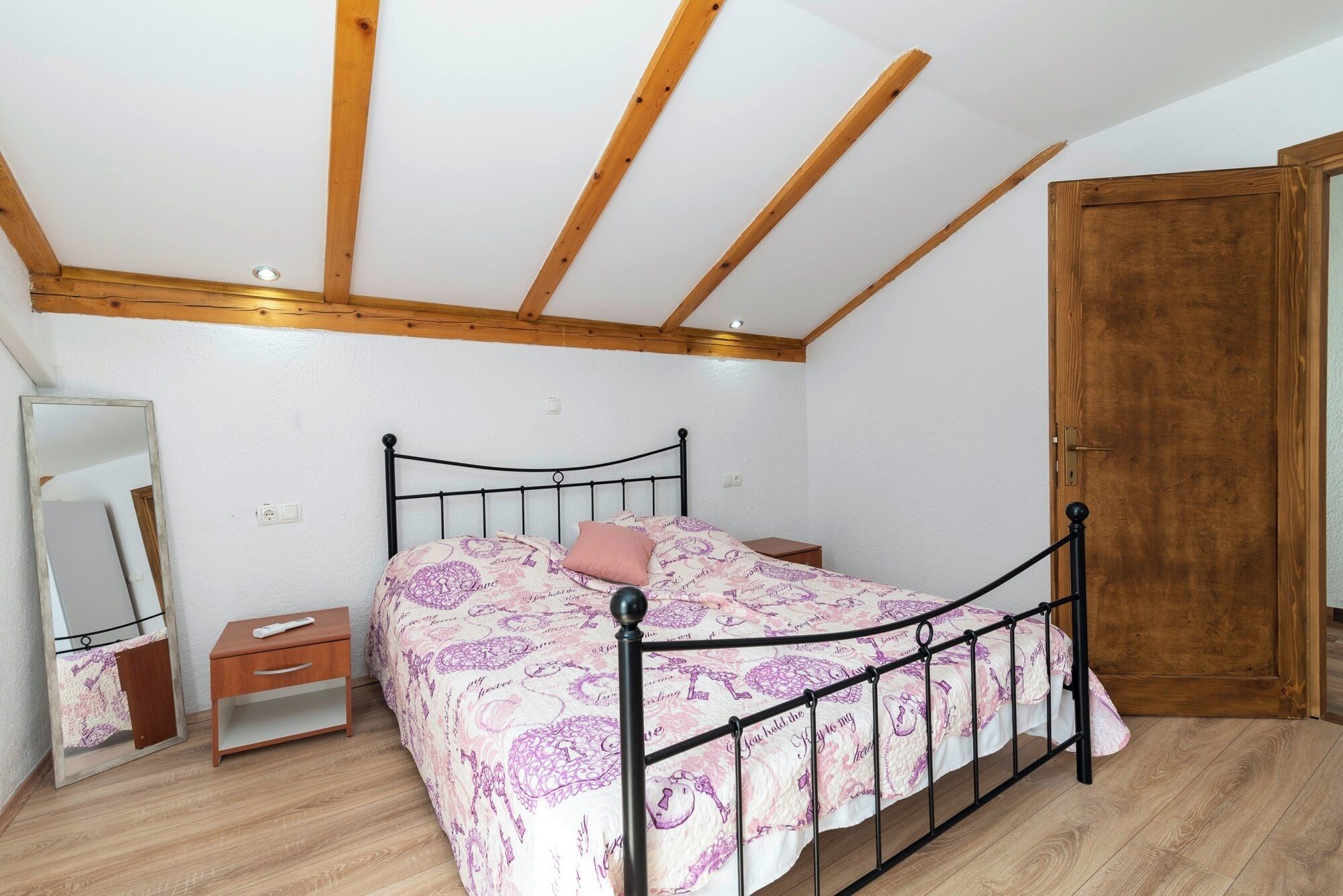 A bedroom with a bed, wooden ceiling beams, a wooden door, a bedside table, a nightstand, and a mirror.