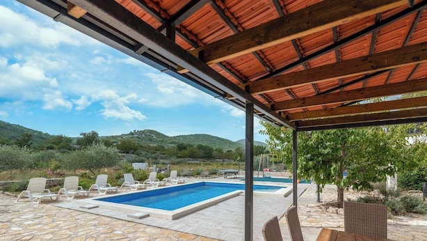 A covered outdoor swimming pool area with stone flooring, lounge chairs, and a pergola with a red roof. - Villa Emanuel in Ljubitovica  - Haus für 4-6 Personen (Ljubitovica)