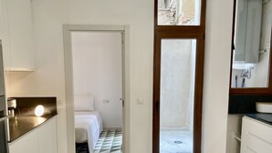 Habitación de hotel con cocina, cama con ropa blanca y baño con espejo y lavamanos.