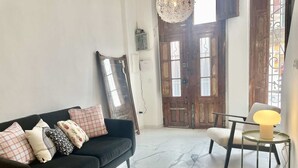 Una sala de estar con un sofá oscuro, cojines estampados, una mesa redonda con una lámpara y una puerta grande de madera con ventanas.