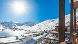 A snow-covered mountainous area with a clear sky and a bright sun. There are buildings with snow on their roofs, and a body of water in the background.