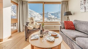 A cozy living room with a large window offering a mountain view, a plush armchair, a sofa, and a coffee table with a bottle, glasses, and a plate.