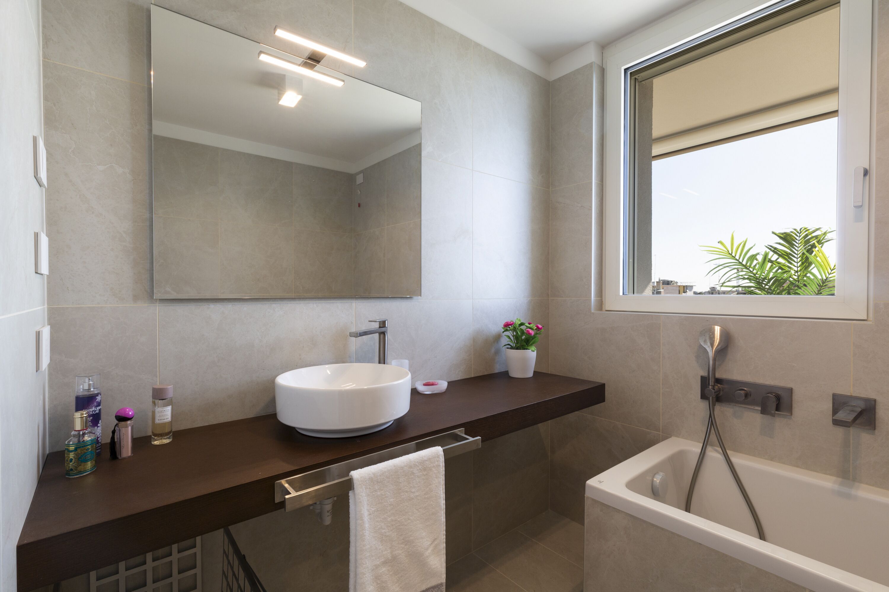 A modern bathroom with a large mirror, a white sink, a bathtub, and a window with a view of greenery.