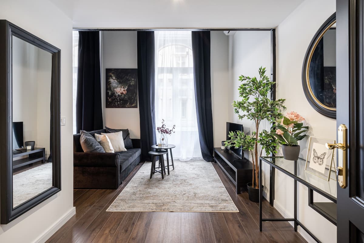 Un salon moderne avec un parquet en bois foncé, un canapé noir, une petite table sur laquelle est posé un vase, et un grand miroir accroché au mur.