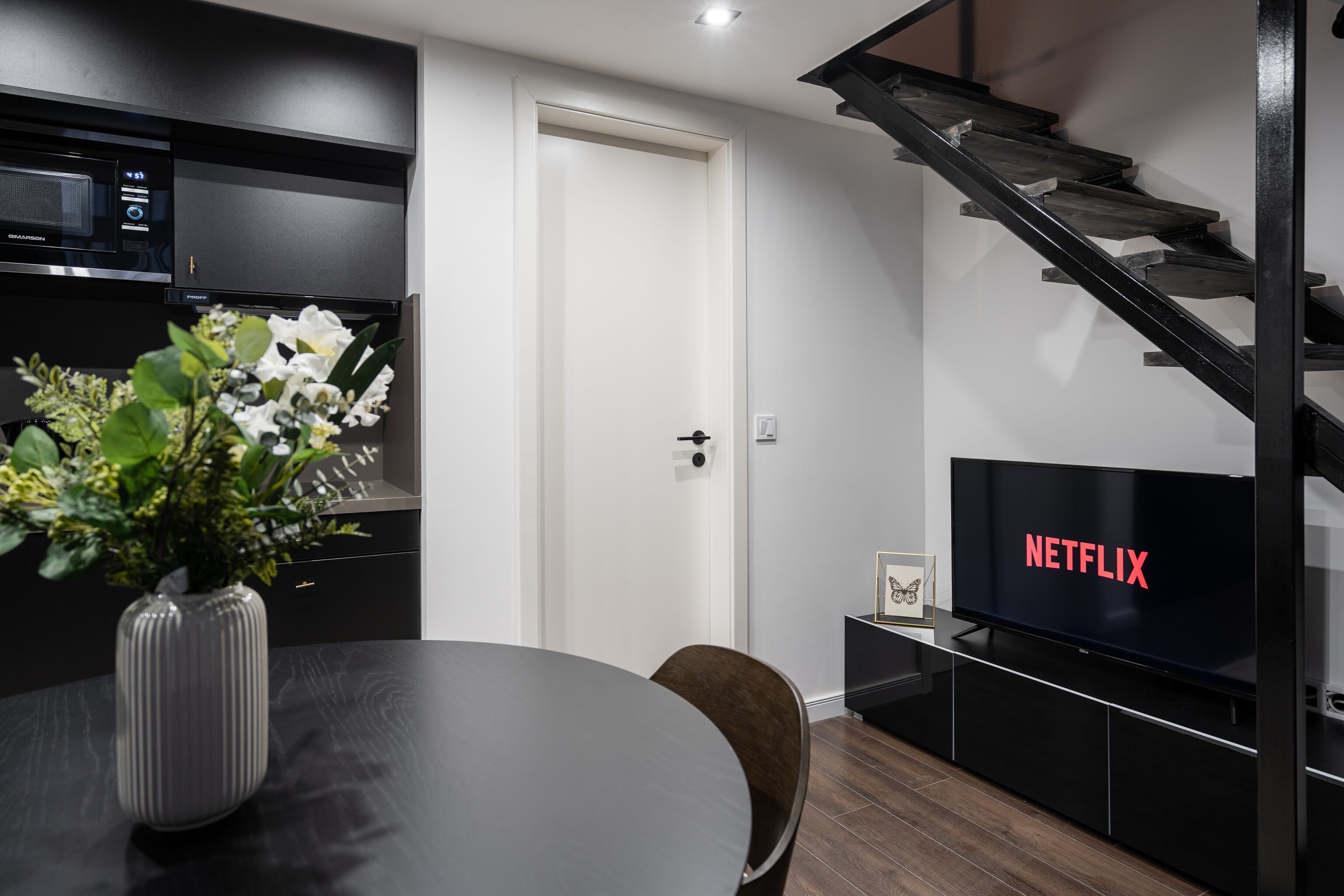 Moderni keittiö, jossa on sisäänrakennettu mikroaaltouuni, ruokapöytä maljakko täynnä kukkia, portaikko ja televisio, jonka näytöllä näkyy Netflixin logo.