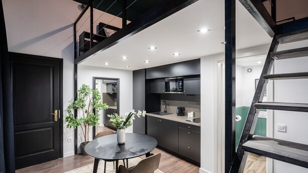 A modern kitchen with dark cabinetry, a black countertop, and a dining area with a round table and chairs. - E41 Boutique Apartments by BQA (Budapest)