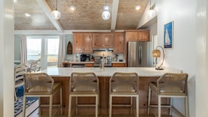 A kitchen with wooden cabinets, a stainless steel refrigerator, and a large island with bar stools. - Feelin' Salty - 107 Quebec 4 Bedroom Home by RedAwning (Dauphin Island)