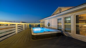 A wooden deck with a hot tub, white railings, and a building with large windows. - Feelin' Salty - 107 Quebec 4 Bedroom Home by RedAwning (Dauphin Island)