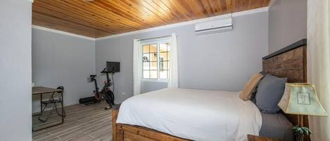 A bedroom with a wooden ceiling, a bed with white bedding, a desk with a chair, and a window.