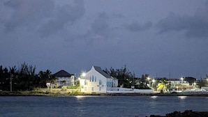 Uma paisagem costeira com uma casa à beira-mar, uma lua crescente no céu e edifícios ao longe.