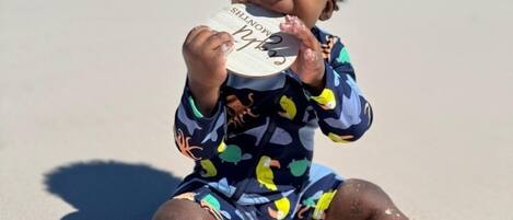 Un niño sentado en una playa arenosa con un cielo azul despejado y el océano al fondo.