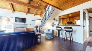 A modern living room with a blue sofa, a stone fireplace, and a staircase leading to a kitchen area with wooden cabinets and a microwave. - Courchevel #5 2 Bedroom Condo by RedAwning (Mammoth Lakes)