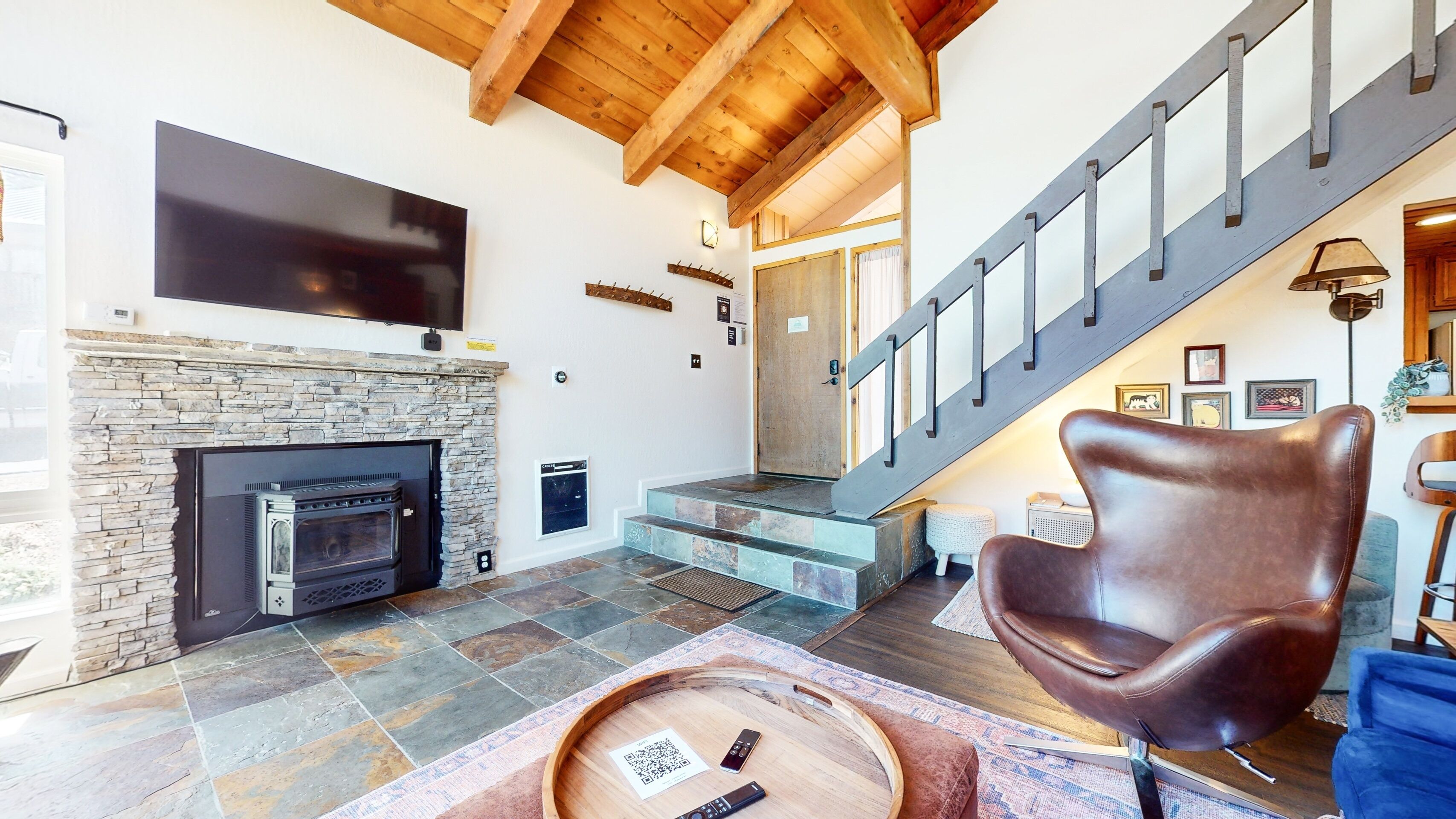A spacious living room with a stone fireplace, a large flat-screen TV, and a wooden staircase.