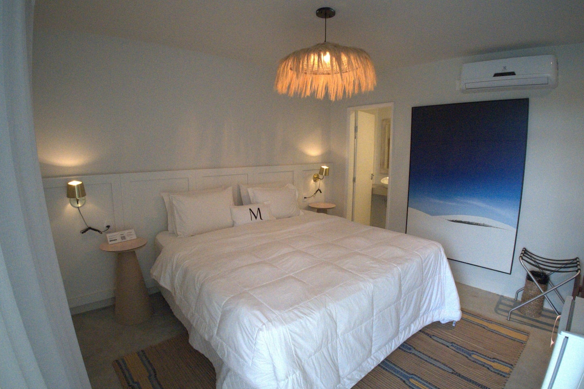 Una habitación de hotel con una cama grande, dos mesitas de noche, una lámpara colgante, un equipo de aire acondicionado y un ventanal con vistas.