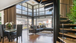 A spacious living area with a large glass wall, a modern sofa, a dining table with chairs, and a wooden staircase. - Always Power SOHO City Retreat Split level Apartment with Table Mountain View (Cape Town)
