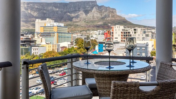 A balcony with wicker furniture, a glass table, and wine glasses. A mountain is visible in the background. - Always Power SOHO City Retreat Split level Apartment with Table Mountain View (Cape Town)