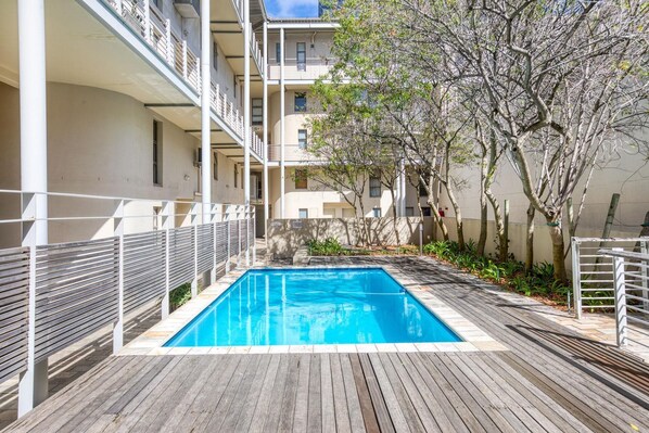 A wooden deck with a rectangular pool, surrounded by a white fence and trees.