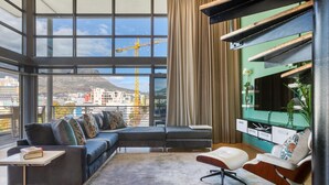 A modern living room with a large window, a sectional sofa, a desk, and a TV mounted on the wall. - Always Power SOHO City Retreat Split level Apartment with Table Mountain View (Cape Town)