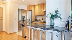 A modern kitchen with wooden cabinets, a stainless steel refrigerator, a microwave, and a coffee machine. There are three bar stools and a vase with flowers on the counter. - Always Power SOHO City Retreat Split level Apartment with Table Mountain View (Cape Town)
