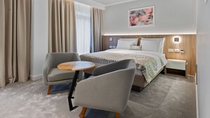 Una habitación de hotel moderna con una cama grande, dos sillones, una mesita redonda y una mesita de noche con lámpara.